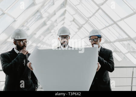 Junge ernster Mann in Helm und Riegel mit Tablet-PC mit zwei Ingenieure begleitet und zeigt auf Gebäude Rohrleitungen. Controller prüfen neue Gebäude. Beratungen Stockfoto