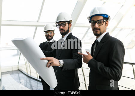 Junge ernster Mann in Helm und Riegel mit Tablet-PC mit zwei Ingenieure begleitet und zeigt auf Gebäude Rohrleitungen. Controller prüfen neue Gebäude. Beratungen Stockfoto