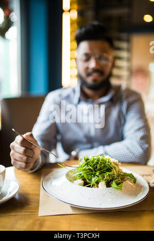 Nahaufnahme von jungen indischen Mann in Hut essen einige köstliche Speisen mit Messer und Gabel im Restaurant. Stilvolle Schüler zu Mittag essen, in der Pause auf un Stockfoto