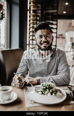 Nahaufnahme von jungen indischen Mann in Hut essen einige köstliche Speisen mit Messer und Gabel im Restaurant. Stilvolle Schüler zu Mittag essen, in der Pause auf un Stockfoto