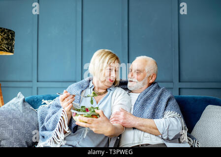 Schönes älteres Paar gesund essen Salat, während mit Plaid auf der Couch zu Hause eingepackt Sitzen Stockfoto