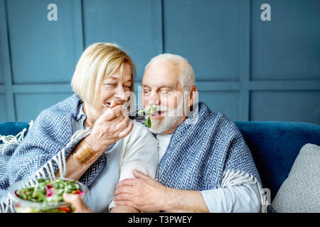 Schönes älteres Paar gesund essen Salat, während mit Plaid auf der Couch zu Hause eingepackt Sitzen Stockfoto