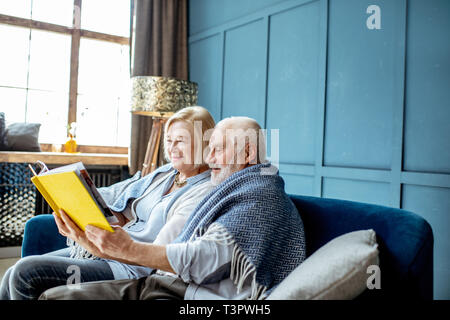 Schönes älteres Paar lesen Buch sitzend mit warmem Plaid auf der Couch zu Hause gewickelt Stockfoto