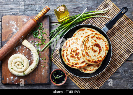 Chinesische Schalotten Pfannkuchen in einer Pfanne mit Ungekochtem Pfannkuchen und Zutaten auf einem Holzbrett, Ansicht von oben, flatlay Stockfoto