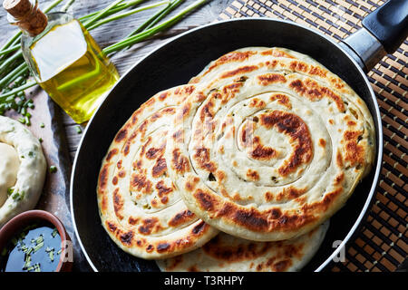 In der Nähe von heißen Chinesischen grüne Zwiebel Pfannkuchen in einer Pfanne mit einem Ungekochten Pfannkuchen und Zutaten auf einem Holzbrett, horizontale Ansicht von oben Stockfoto