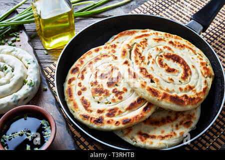 In der Nähe von heißen Chinesischen grüne Zwiebel Pfannkuchen in einer Pfanne mit einem Ungekochten Pfannkuchen, Sesamöl in einer Flasche, Sojasauce und Zutaten auf einem hölzernen Tab Stockfoto