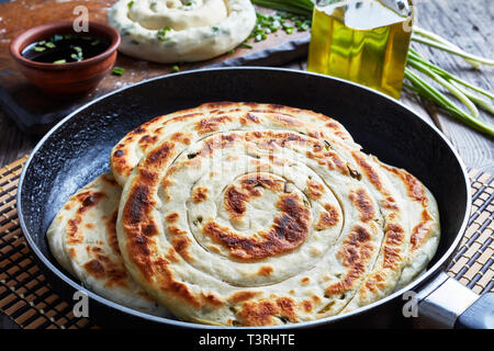 In der Nähe von heißen Chinesischen grüne Zwiebel Pfannkuchen in einer Pfanne mit einem Ungekochten Pfannkuchen, Sesamöl in einer Flasche, Sojasauce und Zutaten auf einem hölzernen Tab Stockfoto