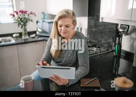Blond tausendjährigen Frau in der Küche sitzen und arbeiten an digitalen Tablet. Arbeiten von zu Hause aus Stockfoto