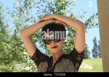 Porträt einer wunderschönen, dunkelhaarige Dame Sonnenbrille tragen in den Park an einem hellen, sonnigen Tag. Hände über dem Kopf. Stockfoto