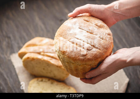 Weibliche Hände, die hausgemachten Naturprodukten frisches Brot mit einer goldenen Kruste auf eine Serviette auf einem alten Holz- Hintergrund. Das Konzept der Backen Backwaren Stockfoto