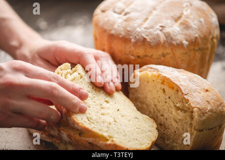 Frauen Hände brechen hausgemachten Naturprodukten frisches Brot mit einer goldenen Kruste auf eine Serviette auf einem alten Holz- Hintergrund. Das Konzept der Backen Backwaren Stockfoto