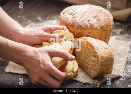 Frauen Hände brechen hausgemachten Naturprodukten frisches Brot mit einer goldenen Kruste auf eine Serviette auf einem alten Holz- Hintergrund. Das Konzept der Backen Backwaren Stockfoto