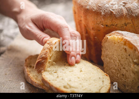 Frauen Hände brechen hausgemachten Naturprodukten frisches Brot mit einer goldenen Kruste auf eine Serviette auf einem alten Holz- Hintergrund. Das Konzept der Backen Backwaren Stockfoto