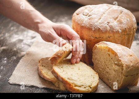 Frauen Hände brechen hausgemachten Naturprodukten frisches Brot mit einer goldenen Kruste auf eine Serviette auf einem alten Holz- Hintergrund. Das Konzept der Backen Backwaren Stockfoto