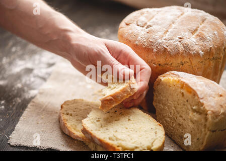 Frauen Hände brechen hausgemachten Naturprodukten frisches Brot mit einer goldenen Kruste auf eine Serviette auf einem alten Holz- Hintergrund. Konzept der Backen Backwaren Stockfoto