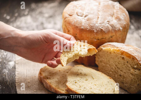 Frauen Hände brechen hausgemachten Naturprodukten frisches Brot mit einer goldenen Kruste auf eine Serviette auf einem alten Holz- Hintergrund. Konzept der Backen Backwaren Stockfoto