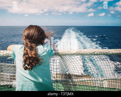 Kleines Mädchen lehnte sich auf das Geländer am Aufwachen durch das Schiff, in dem Sie reist nach links schaut, der Wind bewegt sich ihr Haar und den weißen Schaum der b Stockfoto