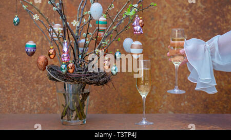 Branchen mit Blüten und Knospen. Ostereier und Osterhasen hängen an den Zweigen. Womans hand Champagner Glas. Festliche Kleidung. Stockfoto