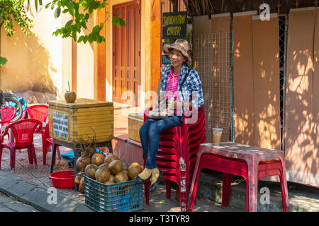 Vietnamesische Frau sitzt außerhalb der Informationszentrale, Hoi An, Quang Nam, Vietnam verkaufen frische Kokosnüsse. Stockfoto