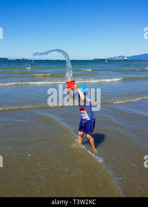 Little Boy spielen im Wasser am Strand. Junge rote Eimer mit Wasser Schlange. Große Schiffe im Hintergrund. Stockfoto