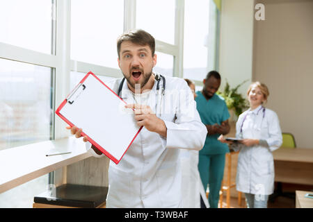 Schön überrascht der männlichen professionellen Arzt stehen über Krankenhaus Hintergrund. Kaukasische Mann mit leeren Banner mit Copyspace. Werbung Bild über das Gesundheitswesen, Gesundheit, Klinik, Medizin. Stockfoto