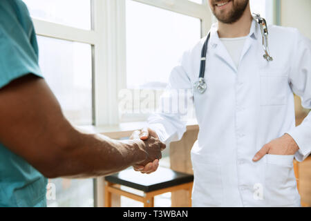 Beschäftigen. Konzept der Zusammenarbeit in der Medizin. Nahaufnahme Foto von zwei Ärzte die Hände schütteln auf grau Krankenhaus Hintergrund. Werbung Bild über das Gesundheitswesen, Gesundheit, Klinik, Medizin und Teamarbeit. Stockfoto