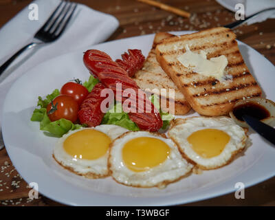 Mehrere Spiegeleier sind auf einer Platte mit Wurst und grünen Salat mit frischen roten Tomaten serviert. Stockfoto