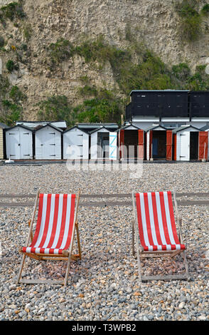 Zwei rot-weiß gestreifte Liegestühle am Strand von Beer in Devon (UK), mit Strandhütten im Hintergrund Stockfoto