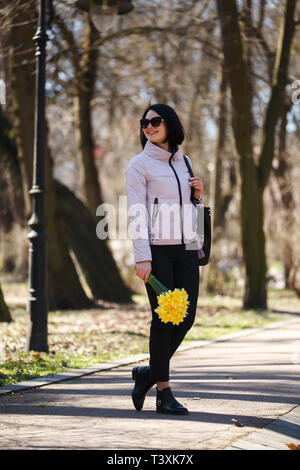 Ein schönes Mädchen in Sonnenbrille hält ein Bündel Narzissen Fuß in den Park Stockfoto