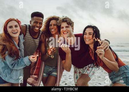 Vielfältige Gruppe von jungen Menschen feiert Tag des neuen Jahres am Strand. Junge Männer und Frauen Spaß mit Wunderkerzen draussen am Meer. Stockfoto