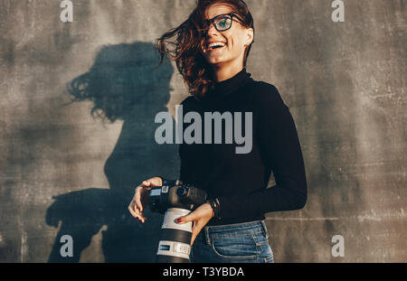 Lachende junge Frau mit einer DSLR-Kamera gegen den braunen Hintergrund. Erfolgreiche Fotografin auf ein Foto schießen. Stockfoto