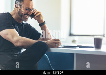 Unternehmer im Gespräch über Handy und der Arbeit an seinem digitalen tablet, während an seinem Schreibtisch sitzen. Geschäftsmann mit Mobiltelefon und lächelnd im Büro Stockfoto