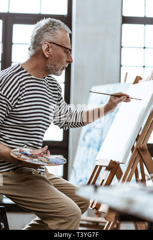 Pinsel in der Hand. Bearded artist in Gläsern holding Malerei Pinsel in der linken Hand, während gemälde landschaft Stockfoto