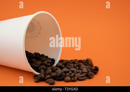 Take-out Kaffeetasse mit dem verschütteten Kaffee Bohnen, orange hinterlegt. Geröstete Kaffeebohnen. Stockfoto