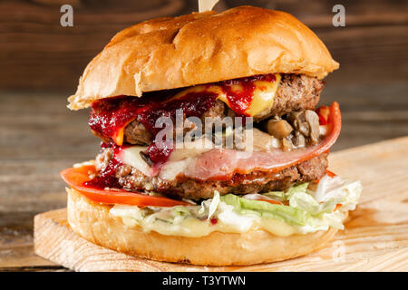 Eine große Burger mit zwei Rind Schnitzel, gekocht auf Holzkohle. Auf einer hölzernen Hintergrund Mittagessen. Das Konzept des Fast Food und nicht Gesund Essen Stockfoto