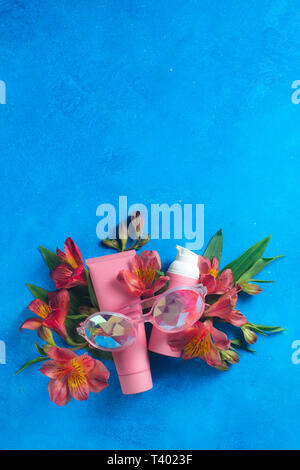 Sonnenbrille und Hautpflege Kosmetik in die rosa Palette in auf einem blauen Hintergrund mit Feder alstroemeria Blumen. Sonnencreme flach mit kopieren. Stockfoto