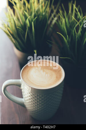 Tasse Cappuccino mit einem Muster auf dem Milchschaum in Form einer Tulpe auf dem Hintergrund der Grünen frische Pflanzen auf einem Holztisch Stockfoto