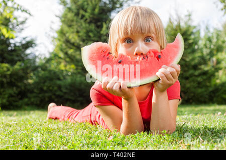 Portrait von elementare Alter Mädchen Spaß beim Essen ein Stück saftige Wassermelone liegen auf einer Wiese im Sommergarten Stockfoto