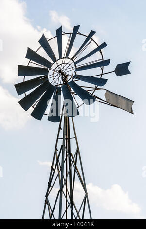 Low Angle View eines altmodischen, multi-Klinge, Metall wind Pumpe auf einem gittermast in der Hintergrundbeleuchtung gegen eine blass blauen Himmel mit weißen Wolken. Stockfoto
