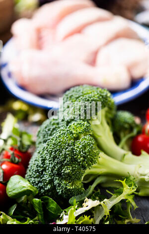 Nahaufnahme von einigen Stämmen von Brokkoli und einige Kirschtomaten auf einem grauen rustikalen Tisch, weiß und blau Keramik Schüssel mit rohem Huhn ich Stockfoto