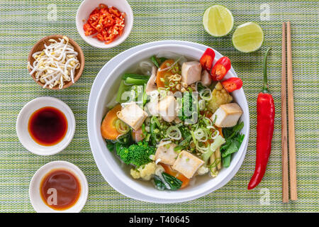 Vegetarische vietnamesische Nudelsuppe Pho diente in Weiße, runde Schüssel, mit Saucen, Kalk, Chili und Sojasprossen. Holz- Stäbchen auf der Seite. Ansicht von oben. Stockfoto