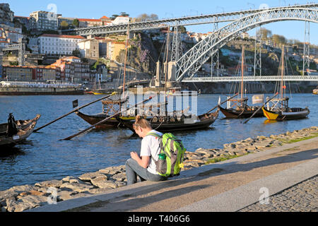 Eine junge Frau sitzt Waterfront riverside Cais de Gaia mit Blick auf Dom Luis Brücke, Fluss Douro und Boote in Porto Portugal Europa KATHY DEWITT Stockfoto