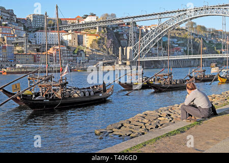 Eine junge Frau sitzt auf der Uferpromenade mit Blick auf Dom Luis I Brücke, Fluss Douro Weingut Boote und Ribeira in Porto Portugal Europa KATHY DEWITT Stockfoto