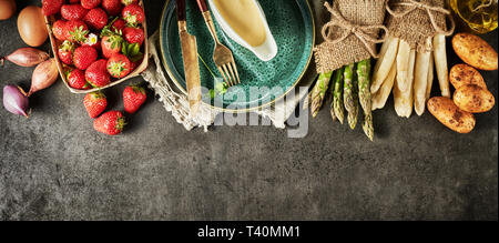 Frischen Zutaten für gourmet Kochen mit Bündel von grünem und weißem Spargel, Erdbeeren, Kartoffeln und Schalotten mit einem leeren Teller ein Stockfoto