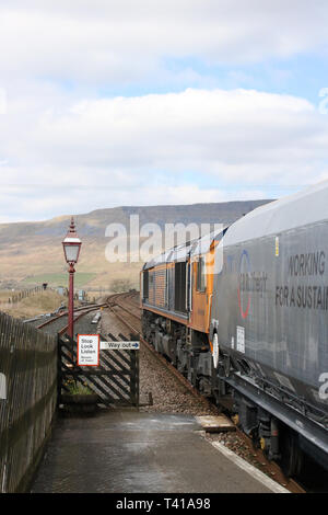 Klasse 66 GBRf diesel-elektrischen Lokomotive schleppen einen Stein Zug durch Ribblehead Station auf der Bahnstrecke nach Carlisle Vereinbaren am 12. April 2019. Stockfoto