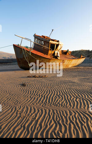 Pwllheli, Gwynedd/Wales - der 10. April 2019: Im Licht der untergehenden Sonne, ein kleiner Rost Boot liegt auf rippled Sand nach den Gezeiten gegangen. Stockfoto