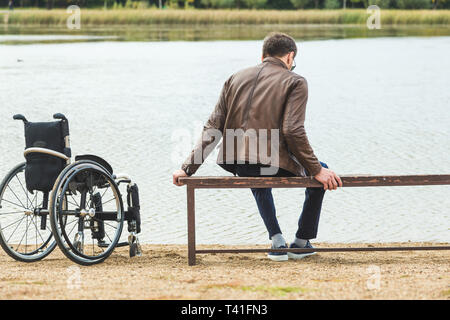 Ein junger Mann sitzt auf einer Bank am See, neben seinem Rollstuhl Stockfoto