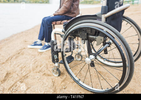 Ein junger Mann sitzt auf einer Bank am See, neben seinem Rollstuhl Stockfoto