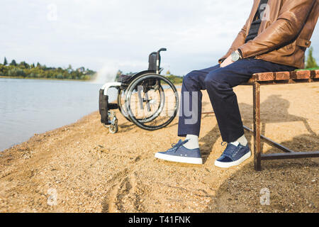 Ein junger Mann sitzt auf einer Bank am See, neben seinem Rollstuhl Stockfoto
