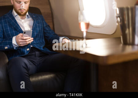 Ein junger erfolgreicher Geschäftsmann in einer teuren Anzug sitzt in den Stuhl eines privaten Jet mit einem Glas Champagner in der Hand und schaut aus dem Fenster. Stockfoto